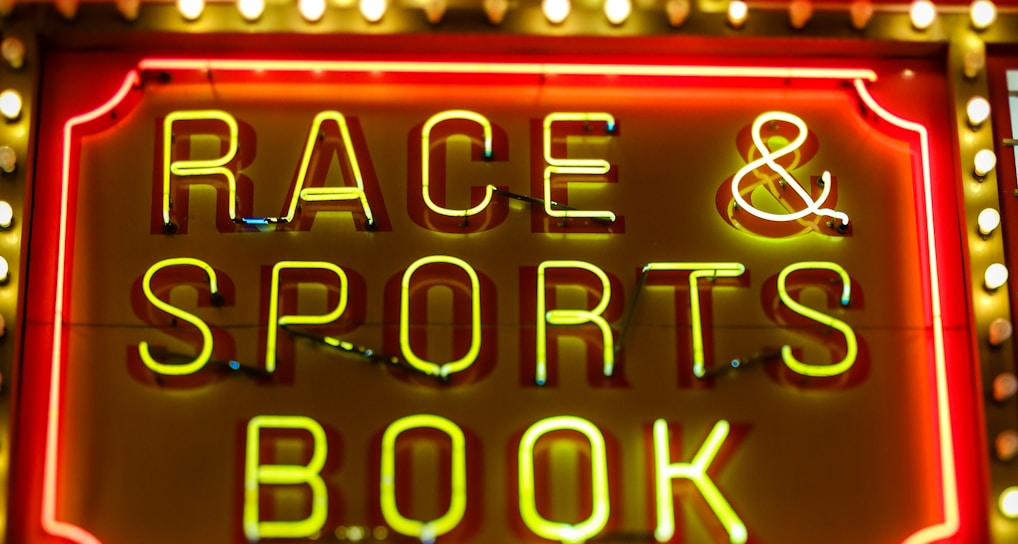 Bright neon lights on a marquee sign display the words 'RACE & SPORTS BOOK'. The sign is surrounded by rows of yellow and white lights against a red background, evoking a vibrant, energetic atmosphere typically associated with casinos or entertainment venues.