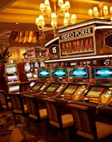 A casino floor featuring rows of brightly lit video poker machines. The area is illuminated by elegant chandeliers, with a display board showing a large sum of money, suggesting a possible jackpot. The rich decor includes luxurious carpets and comfortable seating, adding to the opulent atmosphere.