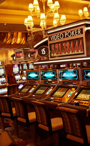 A casino floor featuring rows of brightly lit video poker machines. The area is illuminated by elegant chandeliers, with a display board showing a large sum of money, suggesting a possible jackpot. The rich decor includes luxurious carpets and comfortable seating, adding to the opulent atmosphere.