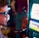 A man wearing sunglasses is intently focused on a slot machine, with colorful lights and graphics illuminating the screen. The scene is set indoors, likely in a casino or gaming area. His attire includes a patterned shirt and a blazer.