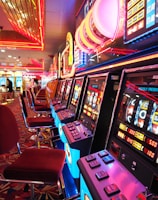 A brightly lit casino interior featuring a row of colorful slot machines. The neon lights create a vibrant atmosphere, with patterned carpeting adding to the decor. Stools are aligned neatly in front of each machine.