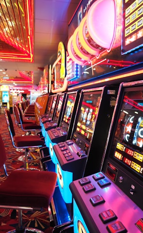 A brightly lit casino interior featuring a row of colorful slot machines. The neon lights create a vibrant atmosphere, with patterned carpeting adding to the decor. Stools are aligned neatly in front of each machine.