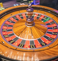 A roulette wheel with alternating red and black numbered pockets on a wooden platform. The wheel contains a small white ball resting in one of the pockets, indicating gameplay. Surrounding the wheel are stacks of colorful poker chips, suggesting a casino environment.