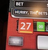 A digital display screen showing a casino betting interface, with text prompting to hurry bets and a highlighted number 27 in red. There are options for betting amounts and a 'share bets' button.