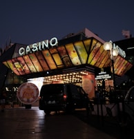 A building with a brightly illuminated entrance is seen at night. The sign above reads 'Casino' in bright letters. The facade features colorful graphics depicting various indulgent themes. Several vehicles are parked near the entrance, and people are visible in the vicinity. Tall lamp posts with spherical lights are present, casting a warm glow.