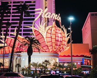 A brightly lit casino with neon lights in vibrant red, pink, and blue, featuring a large flamingo signage. Palm trees are visible in the foreground with vehicles passing by on the street at night.