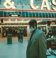 An older man with a gray beard and wearing a suit and hat stands in front of a brightly lit casino. The background features a large illuminated sign reading 'CASINO' and 'SLOTS' among other words, with numerous lights outlining the entrance. Several other people are visible in the background, engaged in various activities, some walking, some standing, and one looking at something on display.